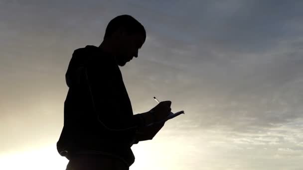 Herrlicher Sonnenuntergang und die Silhouette eines Mannes, der etwas in sein Notizbuch schreibt. — Stockvideo
