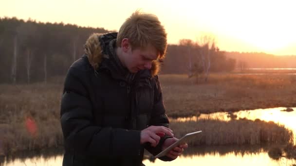 目瞪口呆公平为首的年轻男人正在他平板电脑与触摸屏显示器好日落森林湖与岸上. — 图库视频影像