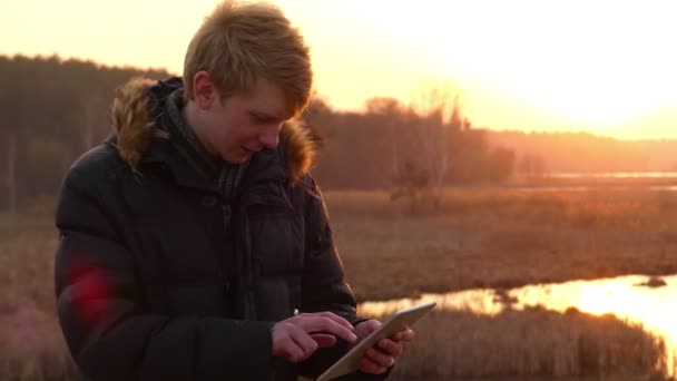 Zaskoczony, młody człowiek pracuje nad jego Tablet komputer z ekranem dotykowym na brzegu lasu jezioro z ładny zachód słońca na wiosnę wczesnego. — Wideo stockowe
