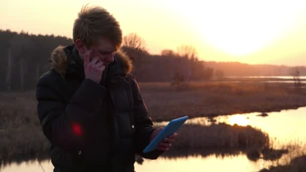 Fair Headed Young Man travaille sur sa tablette avec un écran tactile sur la rive d'un lac forestier avec un beau coucher de soleil au début du printemps . — Video