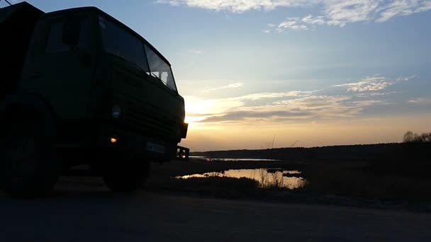 Ein großer LKW fährt bei Sonnenuntergang auf einem Feldweg am See vorbei. — Stockvideo