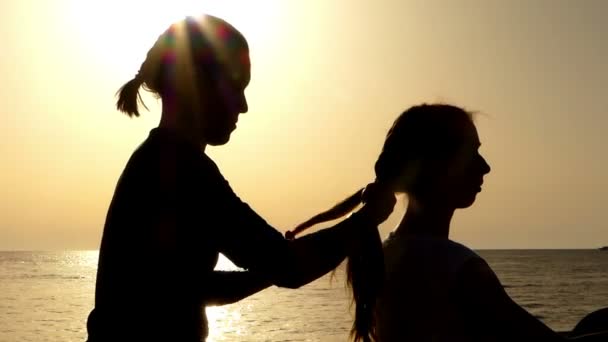 Mutter flechtet Tochter die Haare im Zopf bei Sonnenuntergang am Strand. — Stockvideo