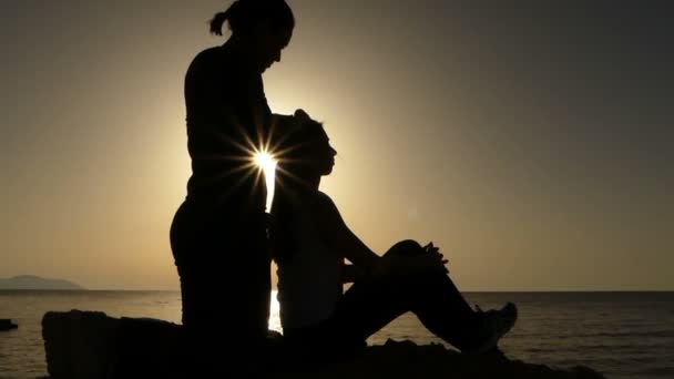 Silhuetas de duas jovens mulheres sentadas e colocando o cabelo em ordem ao pôr do sol . — Vídeo de Stock