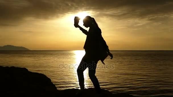 Jeune femme avec une queue de cochon danse sur une plage pierreuse de la mer Rouge, ressemblant à une silouette noire avec un pack dans sa main, au magnifique coucher de soleil — Video