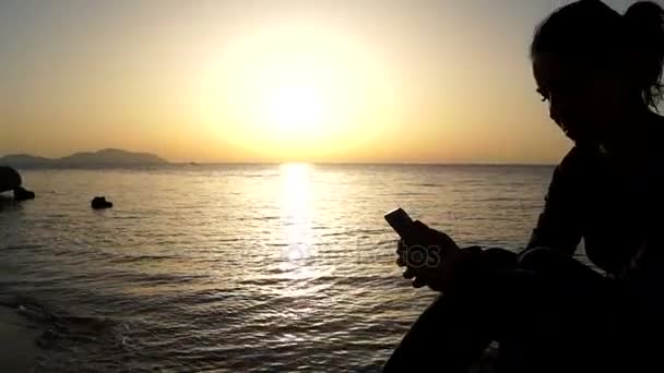Σιλουέτα της γυναίκας με το Smartphone στο χέρι κατά το ηλιοβασίλεμα στην παραλία. — Αρχείο Βίντεο