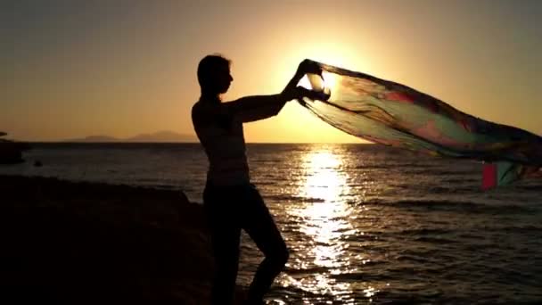 Flickan dansar och leker med sarong i solnedgången på en strand vid havet. — Stockvideo