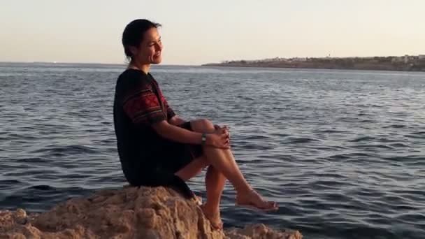 女孩坐着笑对乌克兰穿着石质沙滩上的石头. — 图库视频影像