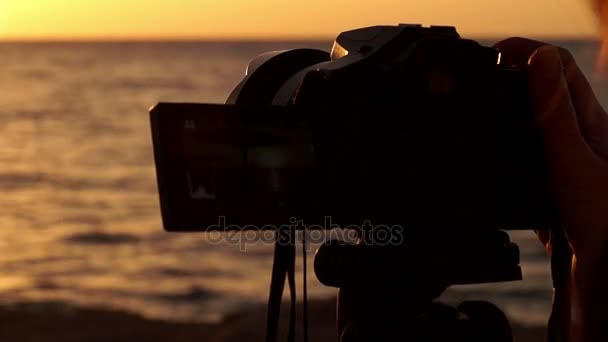 Ruka držící fotoaparát při západu slunce na stativu na pláži u moře.