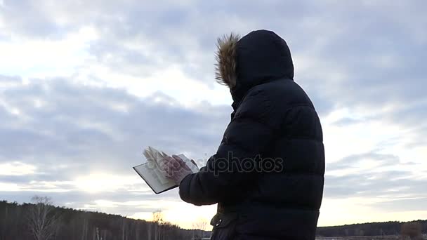Ένας άντρας σε μια κουκούλα στέκεται με την πλάτη στην κάμερα και εξετάζει το βιβλίο κατά το μπλε του ουρανού σε αργή κίνηση. — Αρχείο Βίντεο
