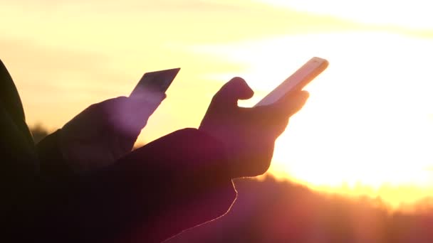 Handen håller kortet och telefonen och personen träder Data från kortet i telefonen på bakgrunden av en gyllene himmel. — Stockvideo