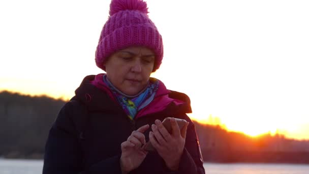 Gyönyörű nő idősebb rózsaszín kalapot néz ki az adatokat egy okostelefon, meglepett, nevet, kint során naplemente. — Stock videók