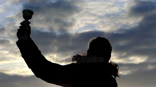 Человек держит трофей и уверенно поднимает его на фоне вечернего неба в медленном движении, его силуэт виден — стоковое видео