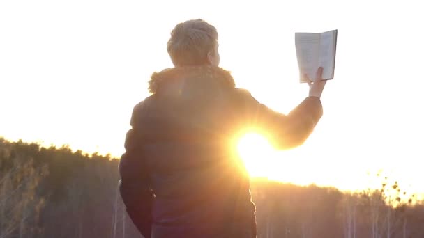 Мальчик читает книгу высоко в руке во время заката в медленном движении — стоковое видео