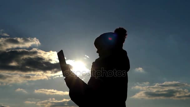 Uma silhueta de uma mulher mulher usa uma tabuleta, a ação na frente do sol e contra o céu azul nublado — Vídeo de Stock