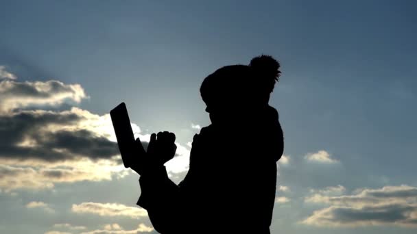 Eine Silhouette einer Frau verwendet ein Tablet, die Aktion vor der Sonne und gegen den blauen bewölkten Himmel — Stockvideo