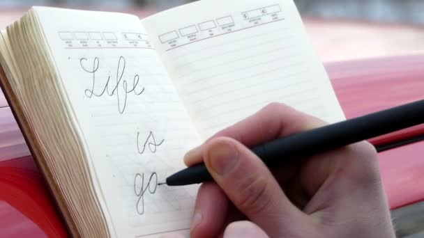 A pessoa no caderno escreve na caligrafia a expressão "a vida é boa" e desenha um sorriso de perto — Vídeo de Stock