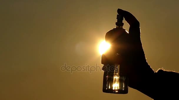 Una persona tiene in mano una bottiglia di profumo e spruzza il liquido sullo sfondo della luce solare e del cielo dorato — Video Stock