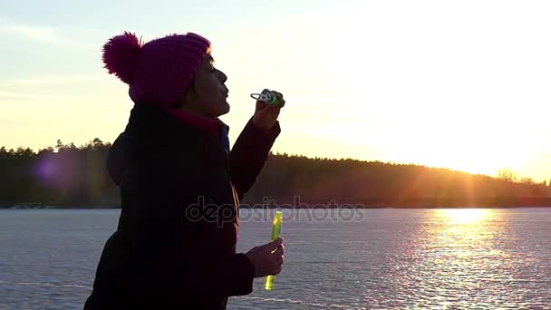 O femeie în creștere umflă bulele de săpun și zâmbește pe fundalul unui lac înghețat în timpul apusului de soare în mișcare lentă — Videoclip de stoc