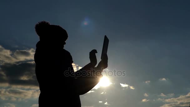 Eine Silhouette einer Frau benutzt eine Tablette und legt sie ab, die Aktion vor der Sonne und vor dem blauen bewölkten Himmel — Stockvideo
