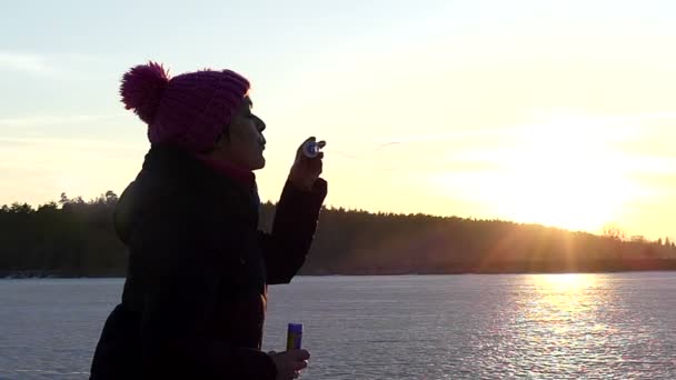 Dospělá žena nafoukne mýdlová bublina na pozadí zamrzlého jezera při západu slunce v pomalém pohybu — Stock video