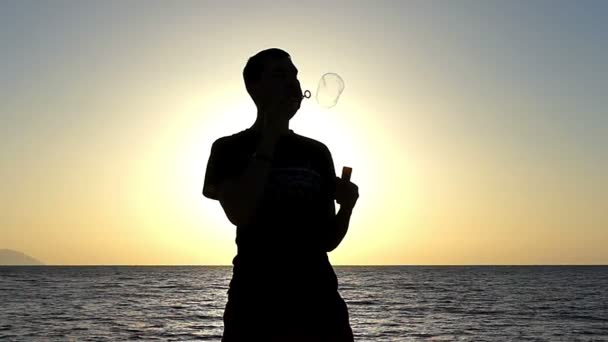Mladý muž dělá mýdlové bubliny na pláži při západu slunce v pomalém pohybu — Stock video