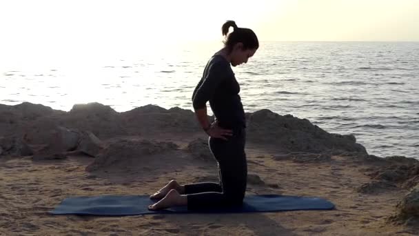 Jeune femme respire profondément à genoux sur un tapis de yoga sur une plage de mer en costume de sport gris — Video