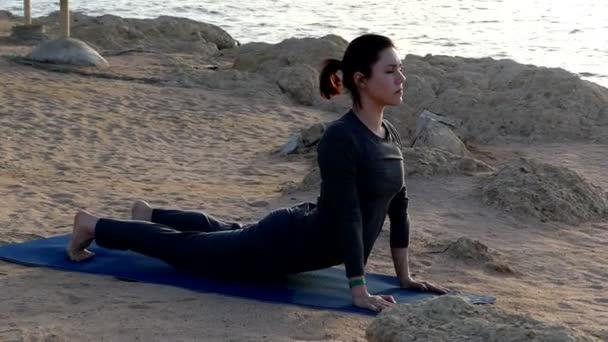 Joven mujer arquea su espalda mientras hace un complejo de yoga en una estera de yoga en una playa de mar en traje deportivo gris — Vídeos de Stock