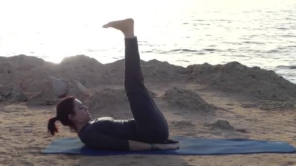 Junge Frau trainiert ihre Bauchmuskeln auf einer Yogamatte und posiert am Strand in einem grauen Anzug — Stockvideo