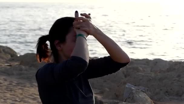 Mujer joven se frota la frente sentada en una esterilla de yoga sobre un fondo marino en traje deportivo gris — Vídeo de stock