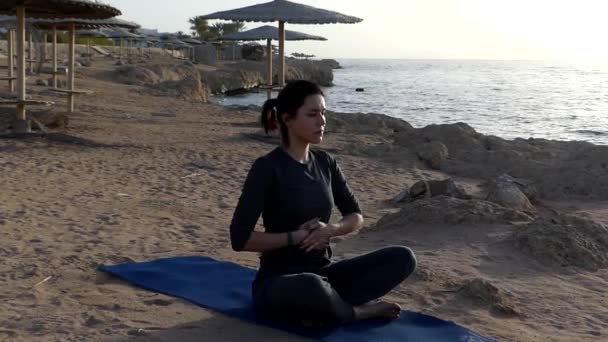 Молодая женщина массирует живот, сидя на коврике для йоги на морском дне в сером спортивном костюме — стоковое видео