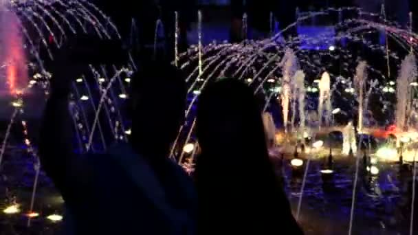Молодой человек и женщина стоят у фантастического фонтана и фотографируются ночью — стоковое видео