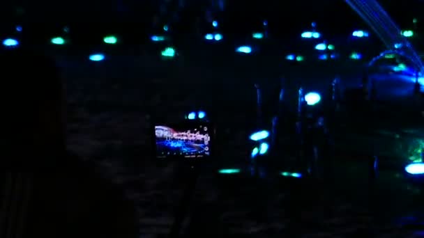 Молодая женщина стоит у фантастического фонтана и стреляет в него мобильным телефоном ночью — стоковое видео