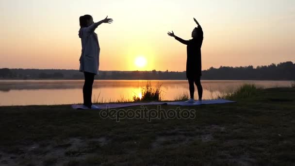 Dos mujeres hacen ejercicios de respiración de yoga al atardecer Neas a un lago en cámara lenta — Vídeo de stock