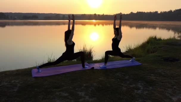 Duas jovens mulheres fazem exercícios de guerreiro de ioga ao pôr do sol em um banco de lago n Slo-Mo — Vídeo de Stock