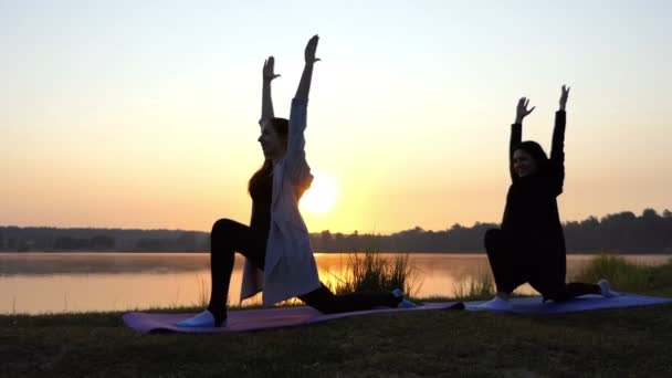 Duas jovens mulheres fazem ioga e sorriem no esplêndido banco do lago ao pôr do sol em Slo-Mo — Vídeo de Stock