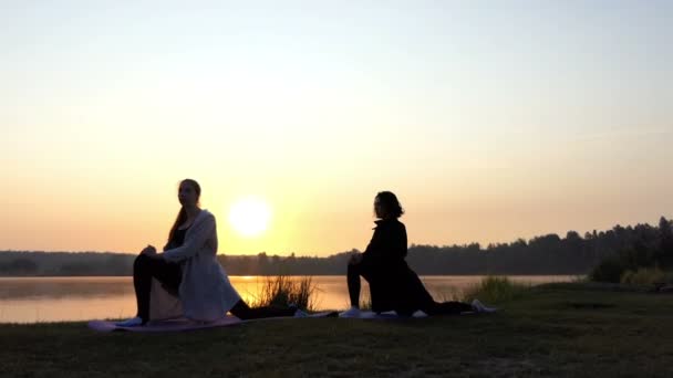 Две девушки стоят на ковриках и растягиваются, делая выпады вперед на закате в Сло-Мо — стоковое видео