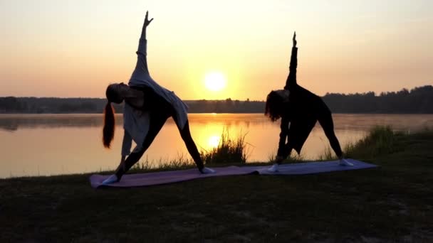 Deux femmes debout sur des tapis de yoga et plier à droite sur une rive du lac au coucher du soleil — Video