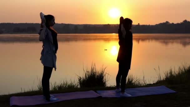 Una chica embarazada hace un ejercicio de yoga con su entrenador en una orilla del lago al atardecer en Slo-Mo — Vídeo de stock