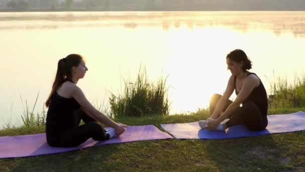 두 젊은 여성이 Slo-Mo에 일몰에 호수 은행에 매트에 앉아 요가 연습 — 비디오