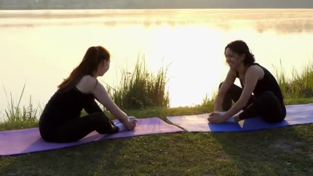 Две женщины сидят на ковриках, наклоняются вперед и держат ноги вместе на закате — стоковое видео