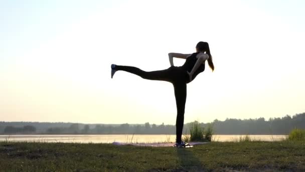 Gravid flicka står på en matta, böjar framåt, på en sjön Bank vid solnedgången — Stockvideo