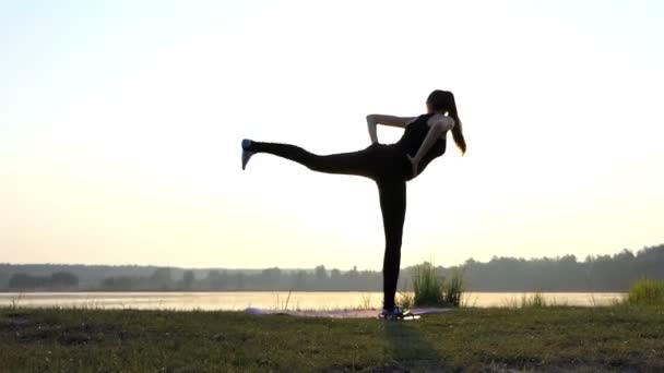 Těhotná dívka stojí na rohoži, ohýbá dopředu, na břehu jezera v západu slunce — Stock video