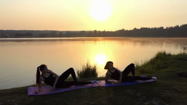 Dos mujeres jóvenes practican yoga acostadas en colchonetas en una orilla del lago al atardecer — Vídeos de Stock