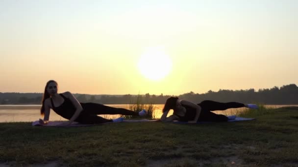 Zwei junge Frauen praktizieren Yoga auf Matten liegend am Seeufer bei Sonnenuntergang im Slo-mo — Stockvideo
