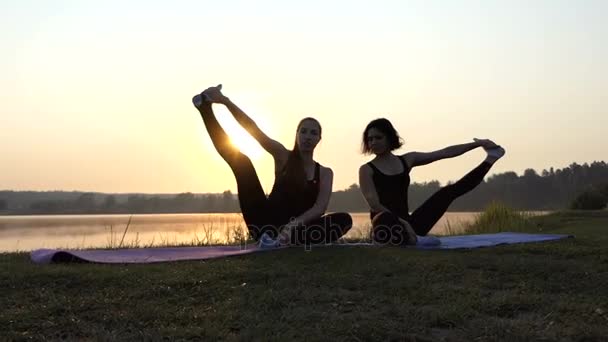 Dos mujeres se sientan en las esteras, levantan las piernas al lado, sonríen, en una puesta de sol espléndida — Vídeos de Stock