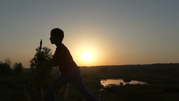 Hits de menino com golpes de Kung-Fu com uma vara em um banco de lago ao pôr do sol — Vídeo de Stock