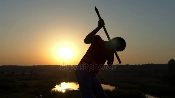Chico entrega golpes de Kung-Fu con un palo en una orilla del lago al atardecer — Vídeos de Stock
