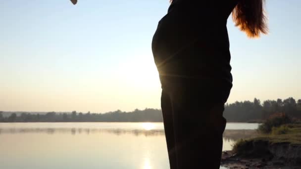 Hamile bir kadın bir bebek elbise karnı üzerinde günbatımında koymak — Stok video