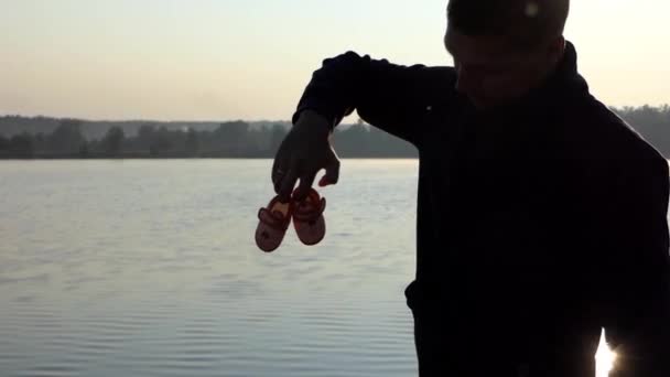 Jovem levanta chinelos de bebê e sorrisos em um banco de lago ao pôr do sol — Vídeo de Stock