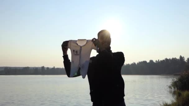 Pai jovem sorri, mantém um terno Romper para seu bebê em um banco de lago ao pôr do sol — Vídeo de Stock